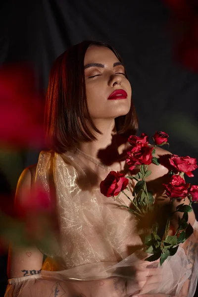 Мрійлива молода жінка в романтичному прозорому платті, що тримає квітучі червоні троянди на чорному тлі — стокове фото