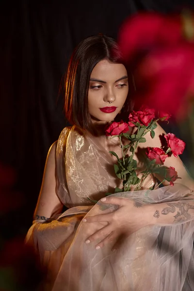 Чувственная молодая женщина в романтическом прозрачном платье держа цветущие красные розы на черном фоне — стоковое фото