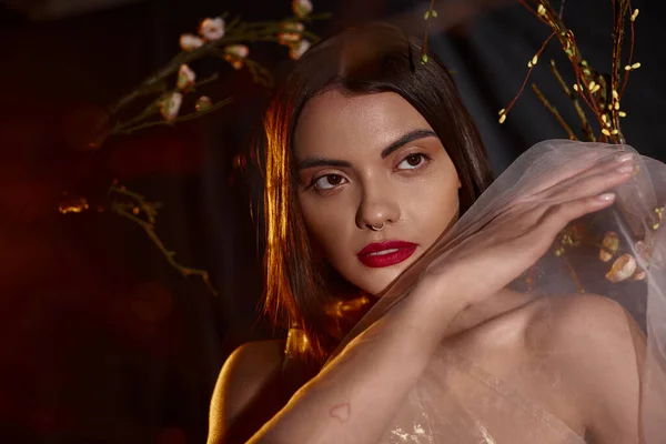 Sensual young woman in transparent dress posing near blooming flowers on branches on black backdrop — Stock Photo