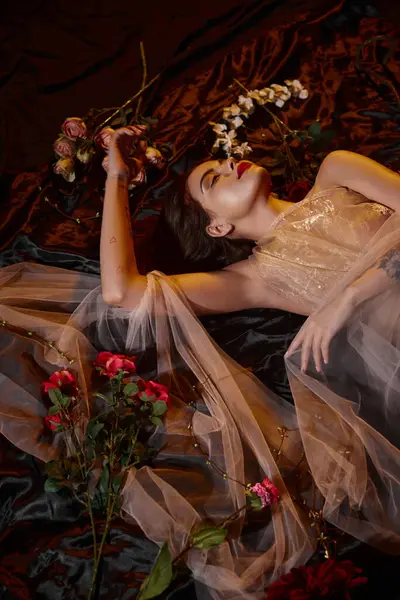Elegant and tattooed young woman in romantic transparent dress lying among blooming flowers — Stock Photo