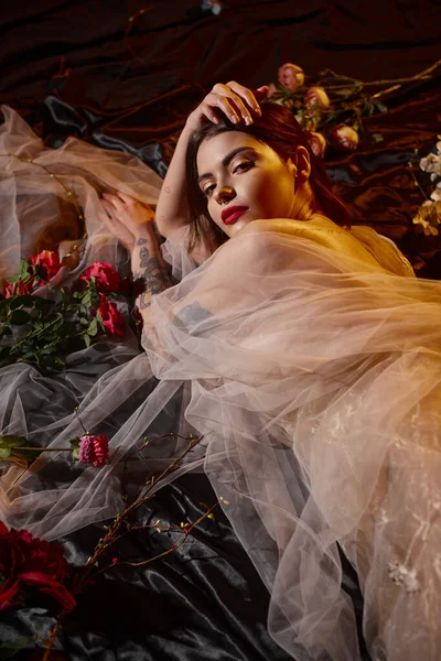 Romantic and tattooed young woman in transparent dress lying among blooming red flowers, elegance — Stock Photo