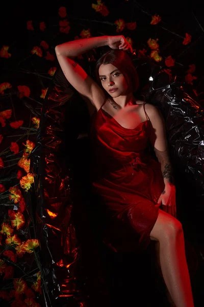 Vista superior, sensual mujer joven en vestido de deslizamiento rojo acostado en bañera negro entre hermosas flores - foto de stock