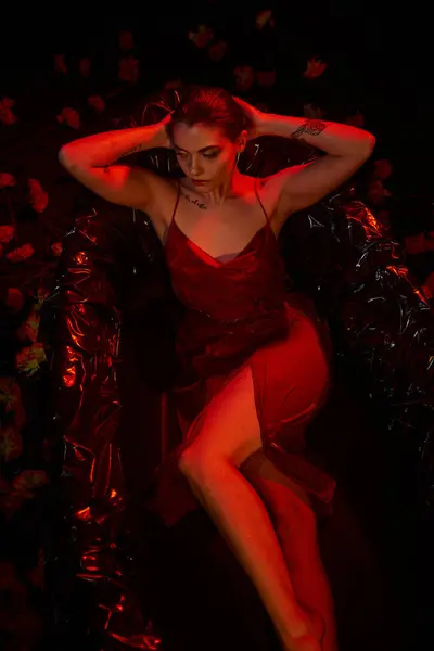 Красный свет, чувственная молодая женщина в скользящем платье, лежащая в черной ванне среди красивых цветов — стоковое фото