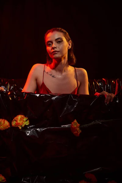 Jeune femme sensuelle en robe glissante mouillée assise dans une baignoire noire parmi de belles fleurs, lumière rouge — Photo de stock