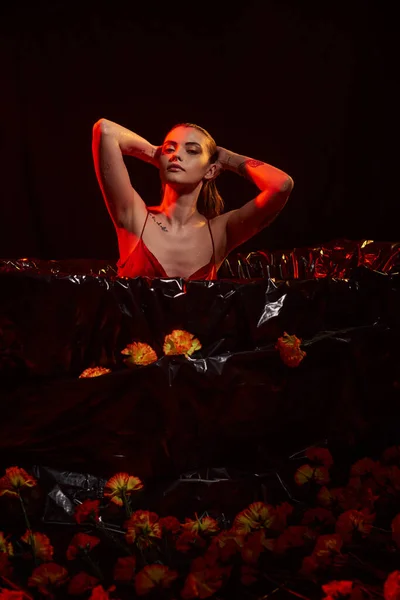 Beautiful young woman in wet slip dress sitting in black bathtub among beautiful flowers, red light — Stock Photo