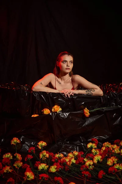 Attractive young woman in wet slip dress sitting in black bathtub among beautiful flowers, red light — Stock Photo