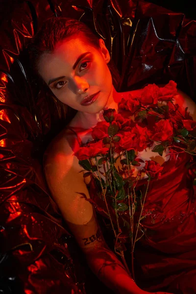 Брюнетка молодая женщина в мокром платье скольжения лежит в черной ванне с цветущими цветами, красный свет — стоковое фото