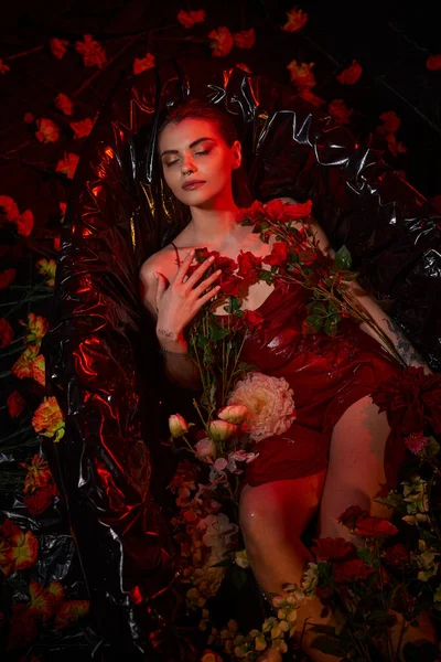 Attractive young woman in wet slip dress sitting in black bathtub with blooming flowers, closed eyes — Stock Photo