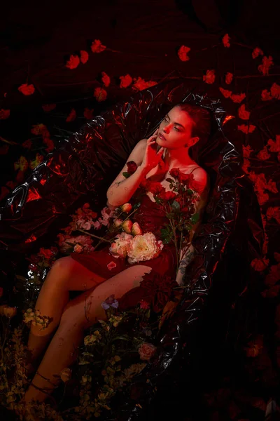 Мрійлива молода жінка в мокрій сукні, що сидить у чорній ванні з квітами, червоне світло — стокове фото