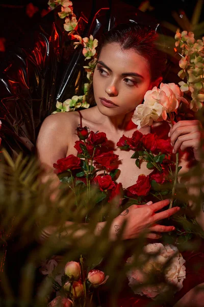 Attractive young woman in wet red dress lying in black bathtub among blooming flowers — Stock Photo