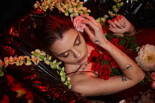 Attractive young woman in wet red dress lying in black bathtub among blooming flowers, top view — Stock Photo