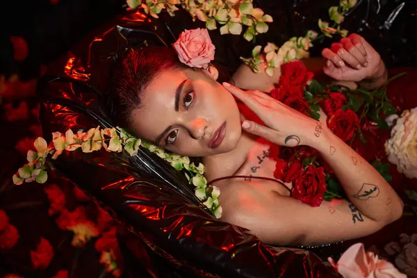 Atractiva mujer en vestido rojo mojado mirando a la cámara y acostado en la bañera entre flores florecientes - foto de stock