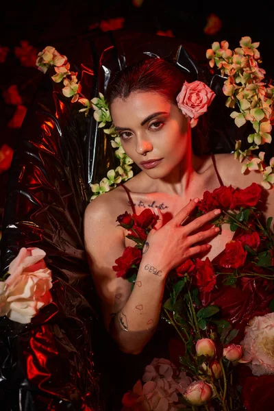 Mulher tatuada em vestido vermelho molhado olhando para a câmera e deitado na banheira entre flores florescendo — Fotografia de Stock