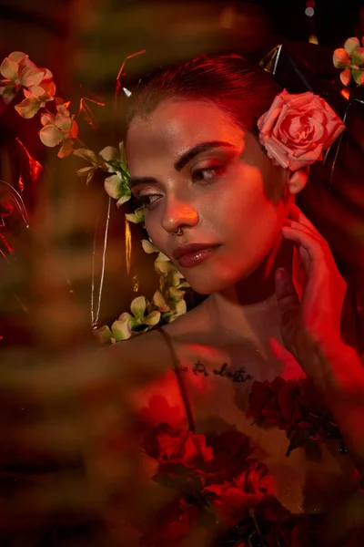 Luz roja en la cara de la joven soñadora con piercing y tatuaje acostado entre diferentes flores - foto de stock