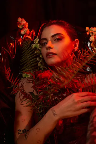 Rotlicht im Gesicht der schönen jungen Frau mit Piercing und Tätowierung, die zwischen verschiedenen Blumen liegt — Stockfoto