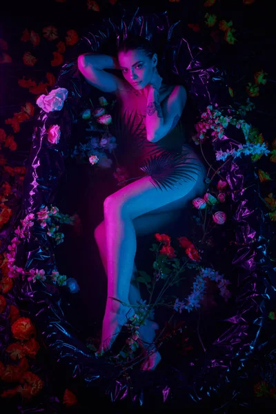 Belleza femenina, mujer joven acostada entre hojas de palma y flores en bañera, luces azules y púrpuras - foto de stock