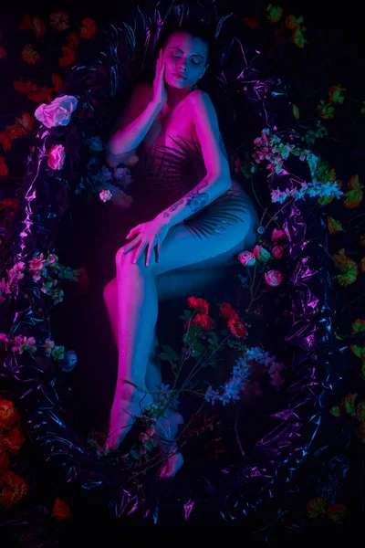 Belleza femenina, mujer bonita acostada entre hojas de palma y flores en bañera, luces azules y púrpuras - foto de stock