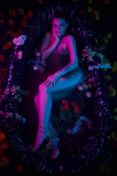 Gracia femenina, mujer bonita acostada entre hojas de palma y flores en bañera, luces azules y púrpuras - foto de stock