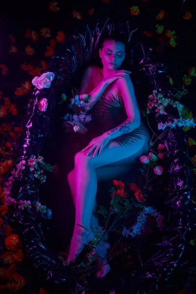 Модний знімок, молода жінка лежить серед пальмового листя і квітів у ванній, синій і фіолетовий вогні — стокове фото