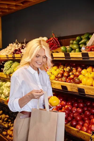 Lustige reife Frau in Freizeitkleidung, die im Supermarkt gelbe Tomaten in die Einkaufstasche steckt — Stockfoto