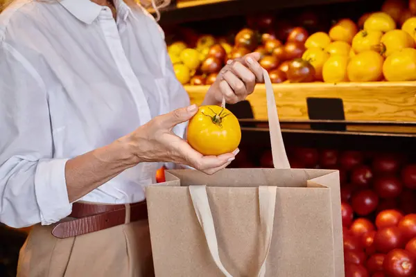 Vista recortada de la mujer madura poniendo tomate amarillo en la bolsa de compras, mientras que en la tienda de comestibles - foto de stock