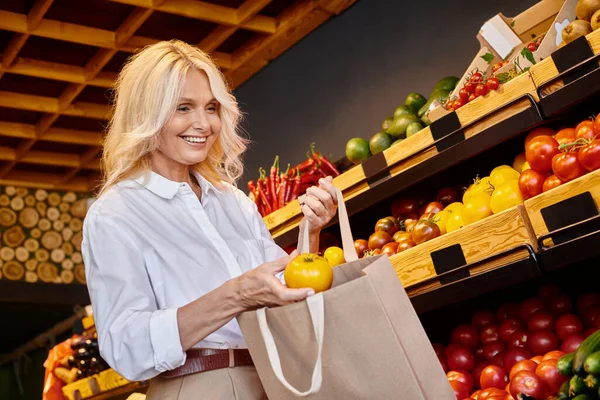 Щаслива зріла жінка в повсякденному вбранні додає жовтий помідор у свою торгову сумку в продуктовому магазині — стокове фото