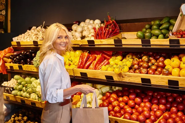 Alegre mujer madura en traje casual sonriendo a la cámara con puesto de verduras en el fondo - foto de stock