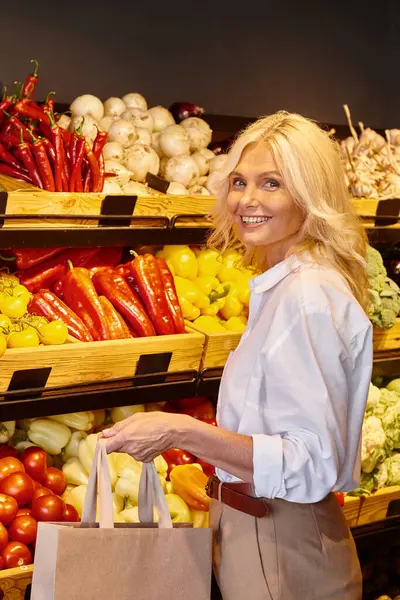 Tiro vertical de la mujer madura con las compras de vuelta sonriendo a la cámara con puesto de verduras en el telón de fondo - foto de stock