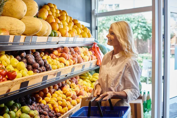 Atractiva mujer madura en ropa casual con bolsa de compras en las manos mirando alegremente a las frutas - foto de stock