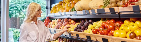 Mujer madura alegre con el pelo rubio en traje casual elegir frutas en la tienda de comestibles, bandera - foto de stock