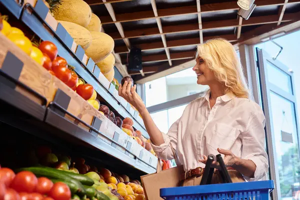 Happy mature woman in casual attire with shopping basket in hands choosing fruits at grocery store — Stock Photo