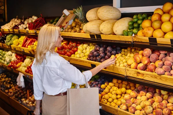 Joyful mature joyous woman in everyday clothes with shopping bag choosing fruits at grocery store — Stock Photo