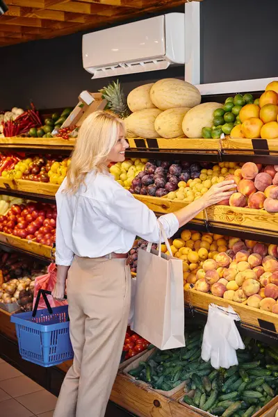 Atractiva mujer madura con cabello rubio con bolsa de compras y cesta en las manos eligiendo frutas - foto de stock