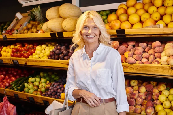 Cheerful mature woman with shopping bag smiling happily at camera with grocery stall on backdrop — Stock Photo