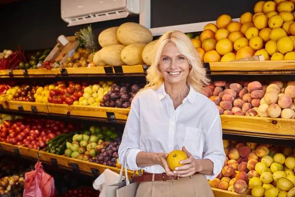 Attractive joyful woman in casual attire posing with orange in hands and smiling happily at camera — Stock Photo