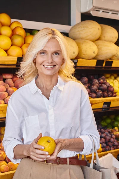 Tiro vertical de mujer feliz posando con naranja y sonriendo a la cámara con tienda de comestibles en el telón de fondo - foto de stock