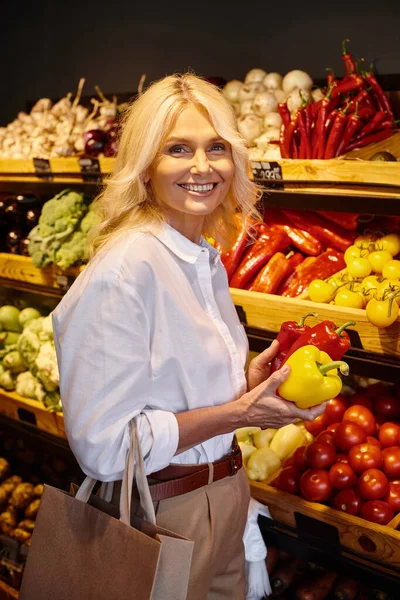 Cheerful mature woman in casual attire holding bell peppers at grocery store and looking at camera — Stock Photo