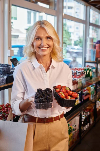 Mujer madura en traje casual posando con fresas y moras en las manos sonriendo a la cámara - foto de stock