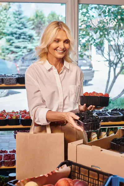 Atractiva mujer madura recogiendo fresas y moras en la tienda de comestibles y sonriendo feliz - foto de stock