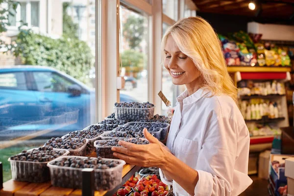 Красивая, зрелая, радостная женщина в повседневной одежде собирает свежие ягоды в продуктовом магазине — стоковое фото