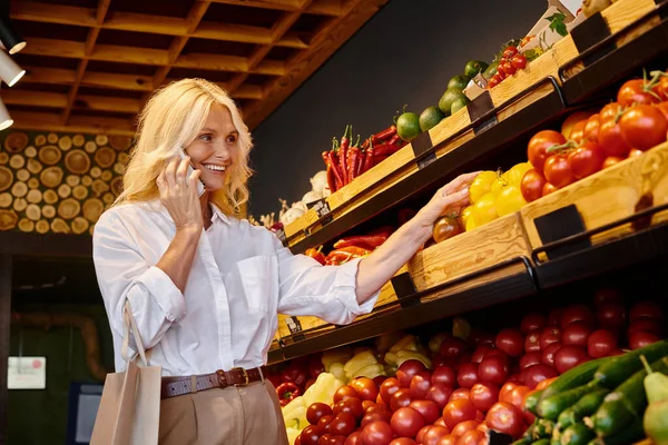 Attractive mature woman picking up vegetables at grocery store and talking joyfully by mobile phone — Stock Photo