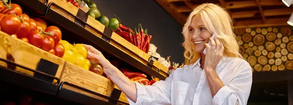 Mujer madura recogiendo verduras en la tienda de comestibles y hablando alegremente por teléfono celular, pancarta - foto de stock