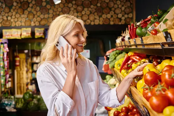 Mujer alegre madura de buen aspecto en traje casual recogiendo verduras y hablando por teléfono móvil - foto de stock