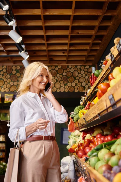 Atractiva mujer madura alegre en ropa casual hablando por teléfono al elegir verduras frescas - foto de stock