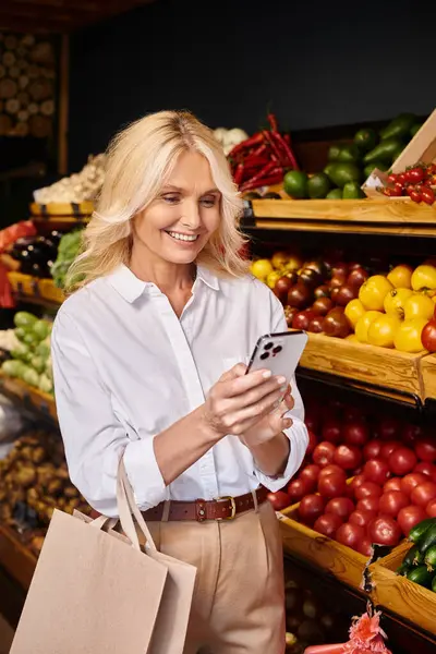 Fröhliche reife Frau in lässiger Kleidung posiert mit Einkaufstasche und blickt glücklich auf ihr Handy — Stockfoto
