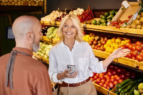 Focus on attractive mature woman asking blurred senior seller about vegetables at his grocery store — Stock Photo