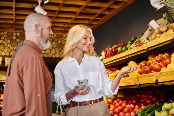 Älterer bärtiger Verkäufer hilft seiner attraktiven Kundin bei der Gemüseauswahl in seinem Lebensmittelladen — Stockfoto