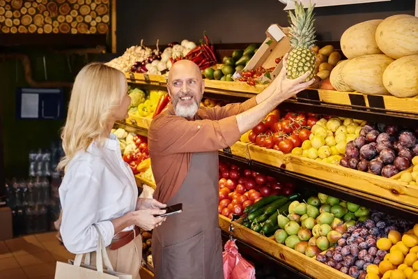 Cheerful bearded seller showing huge pineapple to his female customer next to grocery stall — Stock Photo