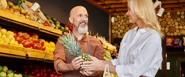 Joyful bearded seller giving fresh pineapple to his mature female customer at grocery store, banner — Stock Photo