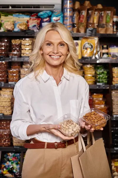 Mujer alegre madura de buen aspecto sosteniendo pistachos y almendras y sonriendo a la cámara alegremente - foto de stock
